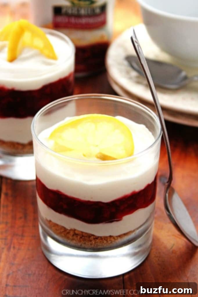 A beautifully layered Lemon Raspberry Cheesecake Trifle in a clear glass, showing distinct layers of graham cracker crust, creamy lemon cheesecake, and bright red raspberry filling. Garnished with fresh lemon slices.