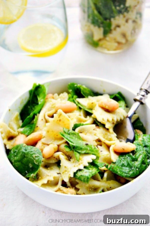 A tantalizing side shot of Pesto Pasta Salad, brimming with bow tie pasta and vibrant spinach, nestled in a pristine white bowl. Garnished with fresh lemon zest and a sprinkle of Parmesan.