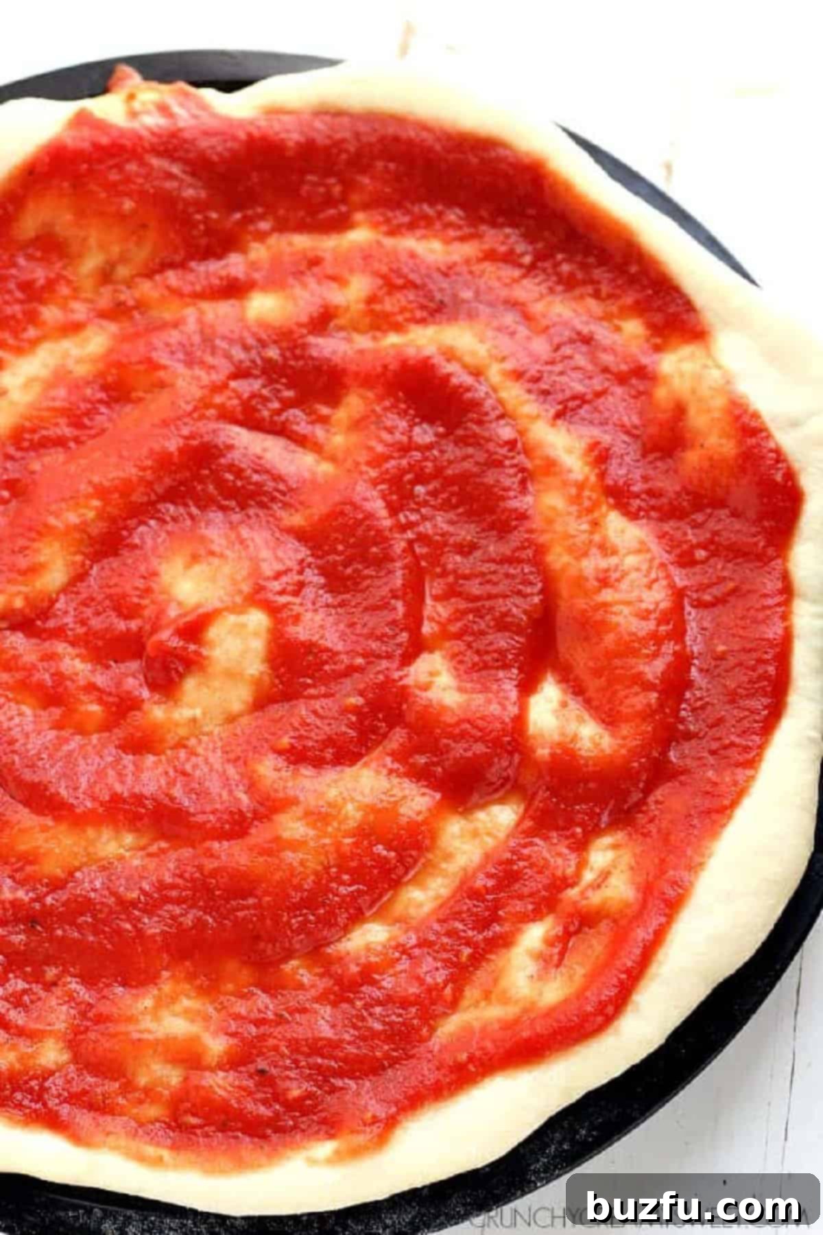 Vibrant red homemade pizza sauce evenly and smoothly spread over a perfectly round pizza dough base, nestled within a baking pan, poised and ready for the addition of delicious toppings.