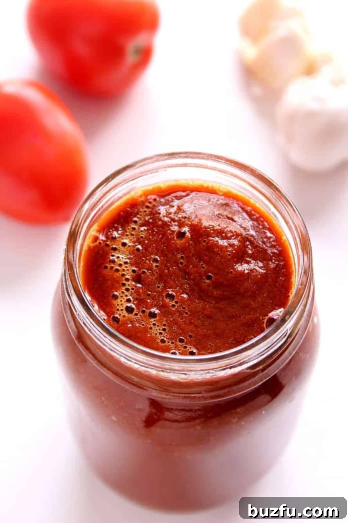 Vibrant red enchilada sauce in a glass jar, perfectly smooth and ready to use. Fresh tomatoes and garlic cloves rest beside it on a white board, highlighting the simple, fresh ingredients that go into this homemade recipe.