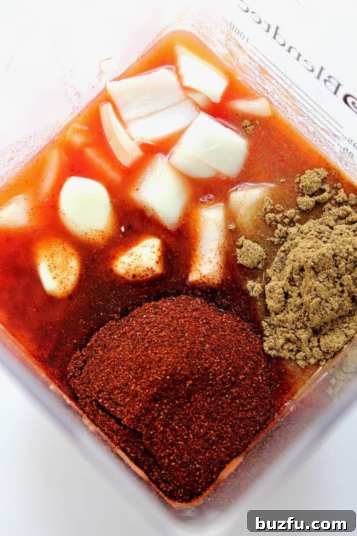 An overhead shot showcasing the key ingredients for homemade enchilada sauce—onion, chili powder, cumin, garlic, and tomato sauce—all gathered inside a blender, ready to be transformed into a delicious sauce.