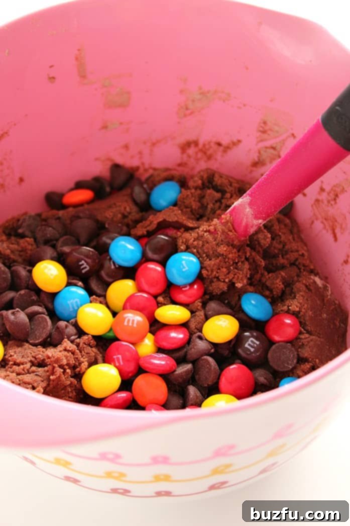 Close-up of chocolate cookie dough generously mixed with colorful M&M's in a mixing bowl.