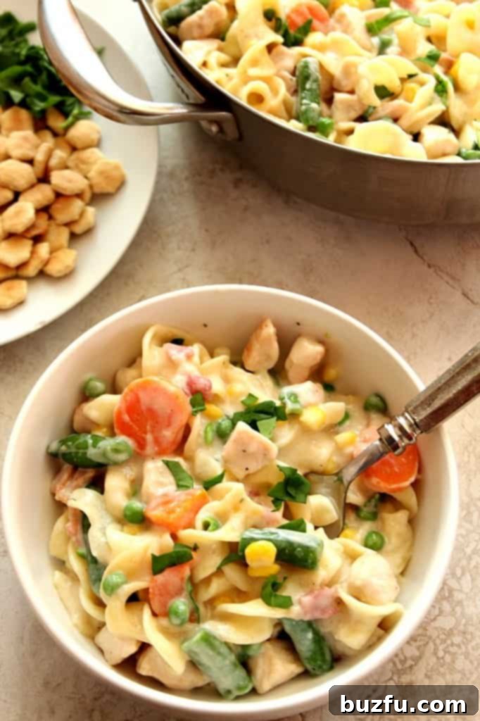 Overhead shot of chicken pot pie pasta in white bowl with skillet in the background, showing the rich, creamy texture and generous chicken pieces.