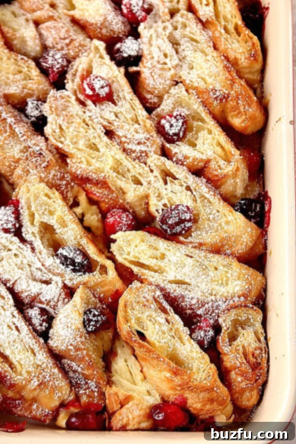 Easy Orange Cranberry Croissant Bake for a festive holiday breakfast or brunch. A golden-brown Orange Cranberry Croissant Bake in a white casserole dish, garnished with powdered sugar and fresh cranberries. Perfect for a holiday breakfast.