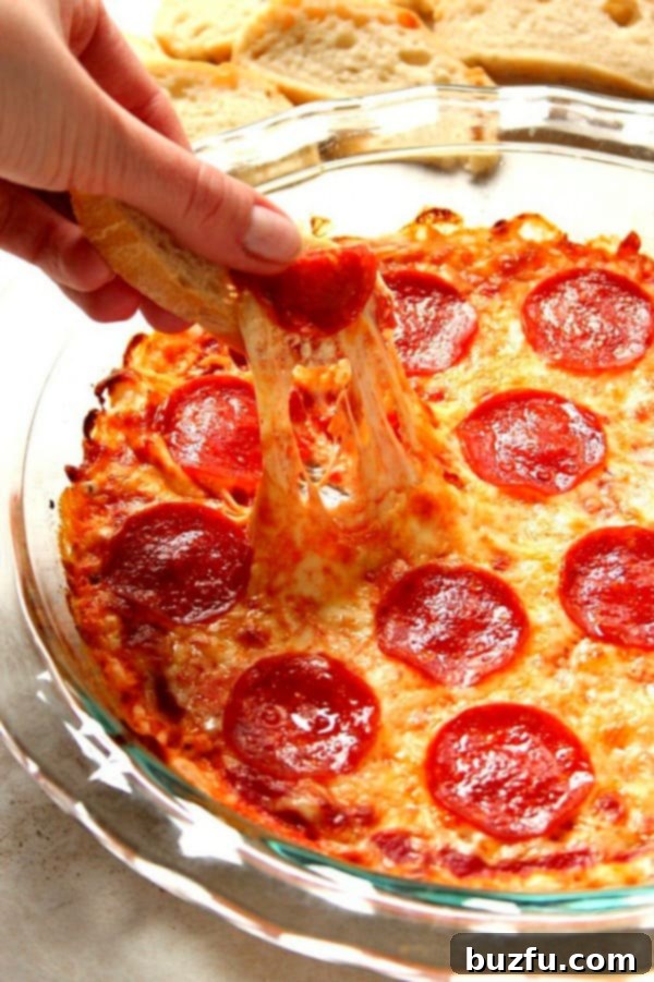 Pepperoni Pizza Dip in glass pie dish with a baguette slice being dipped into the cheesy goodness.