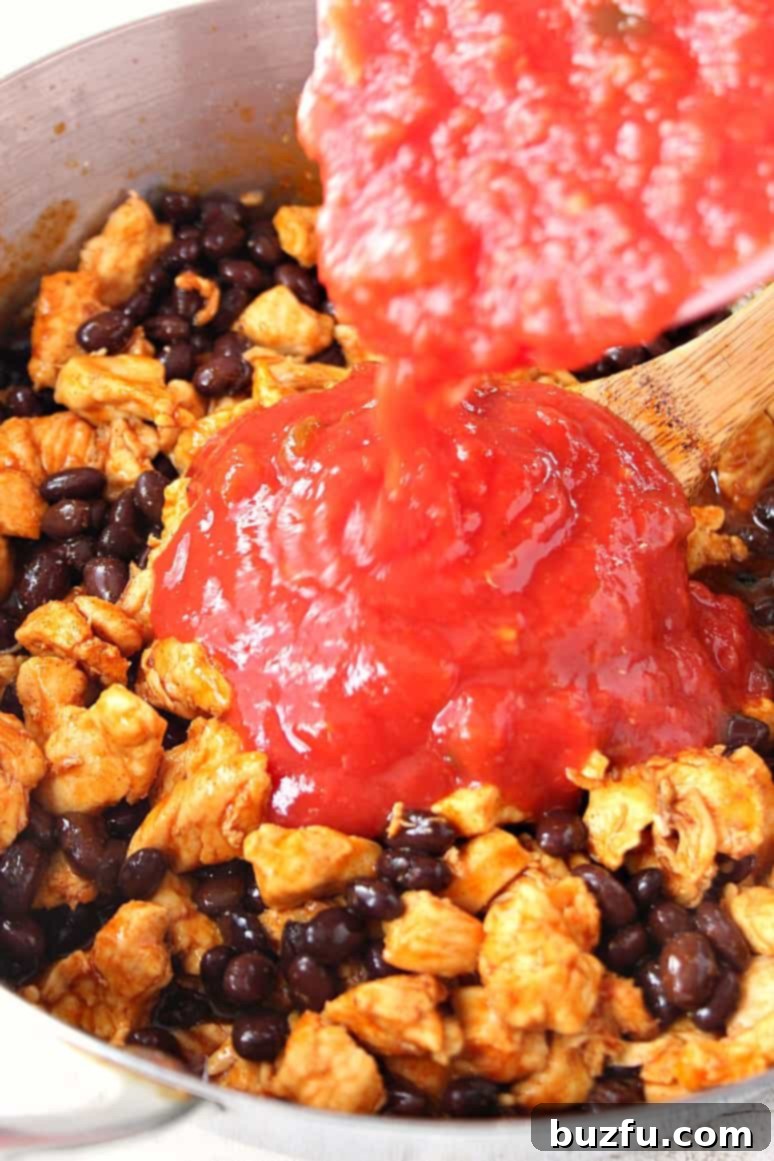 Close-up side view of crushed tomatoes being poured into a skillet, mixing with seasoned chicken and black beans, forming a vibrant sauce.