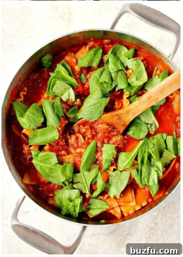 Overhead shot of tomato sauce with sausage, lasagna noodles and fresh spinach on top in skillet, before cheese is added.