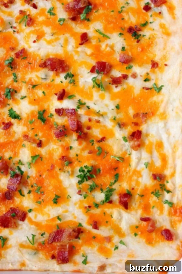 A golden-brown cheesy mashed potato casserole, garnished with melted cheddar, crispy bacon bits, and fresh parsley. Ready to be served in a baking dish.