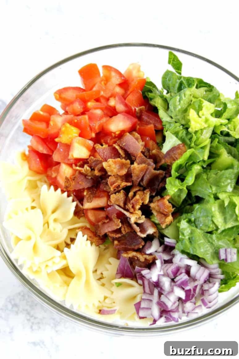 Overhead shot of ingredients for BLT pasta salad in a clear glass bowl, showcasing vibrant lettuce, red tomatoes, and crispy bacon.