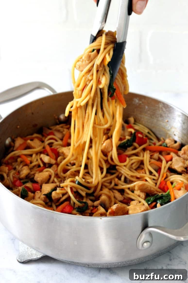 Easy Chicken Lo Mein Recipe - quick and easy homemade chicken, vegetables and noodles dish with teriyaki sauce. Close-up side shot of chicken lo mein being served from a deep skillet with tongs, highlighting the glossy sauce and perfectly cooked ingredients.