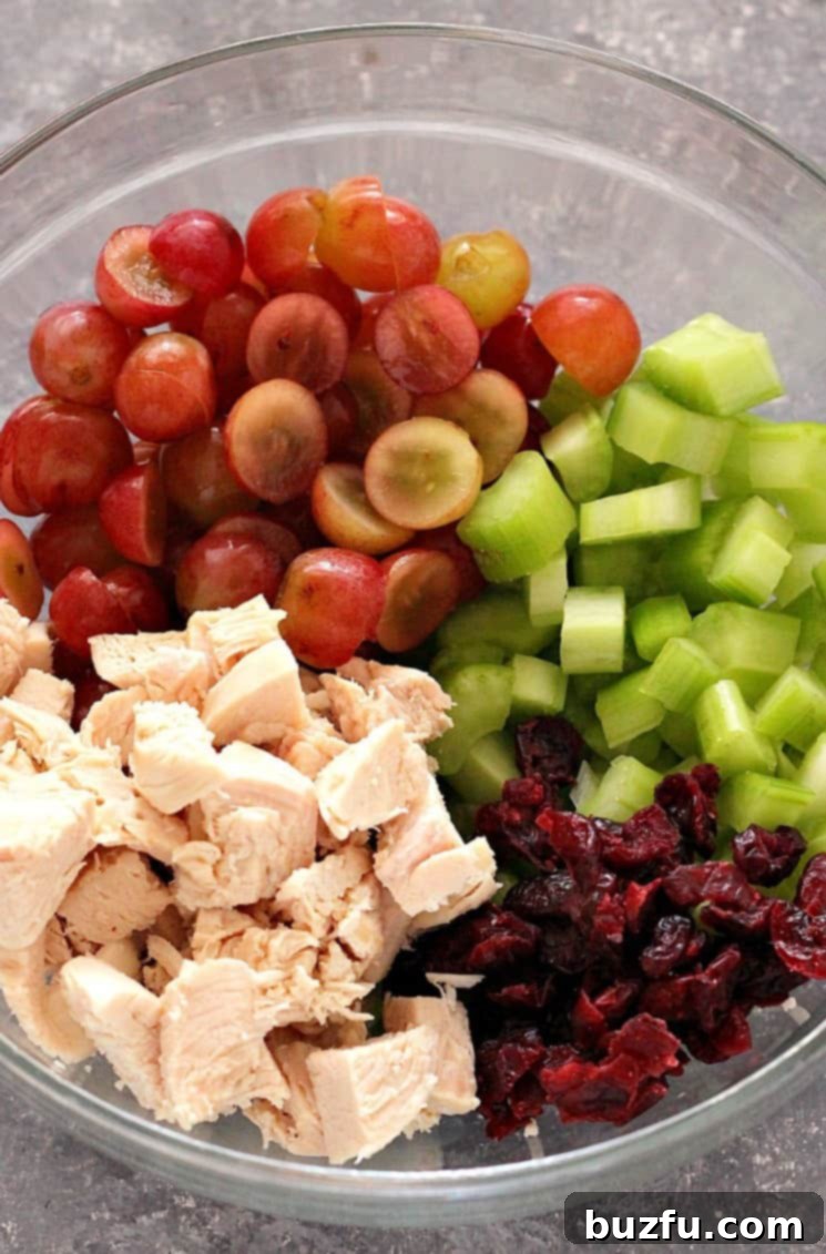 Easy Chicken Salad with Grapes, celery and cranberries, tossed with light sour cream dressing. Perfect lunch you can make ahead and enjoy served in sweet rolls. Overhead shot of cubed chicken, grapes, celery and craisins in glass bowl, ingredients for chicken salad.