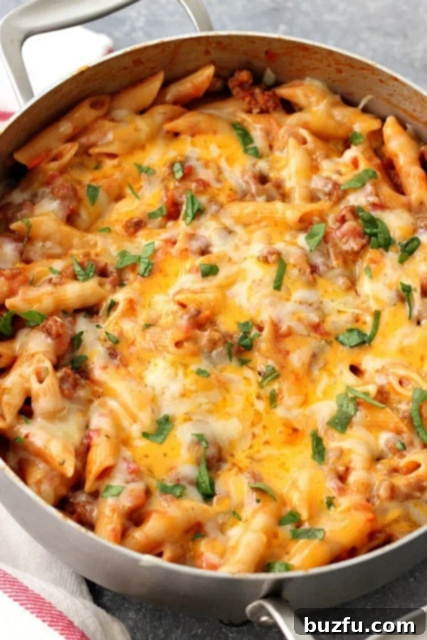 One Pot Cheesy Sausage Penne Recipe - hearty and satisfying one pan pasta dinner. Italian sausage, quick tomato sauce and penne pasta with cheesy topping is perfect for busy weeknights. A large skillet filled with creamy, cheesy one-pot sausage penne pasta, garnished with fresh herbs.