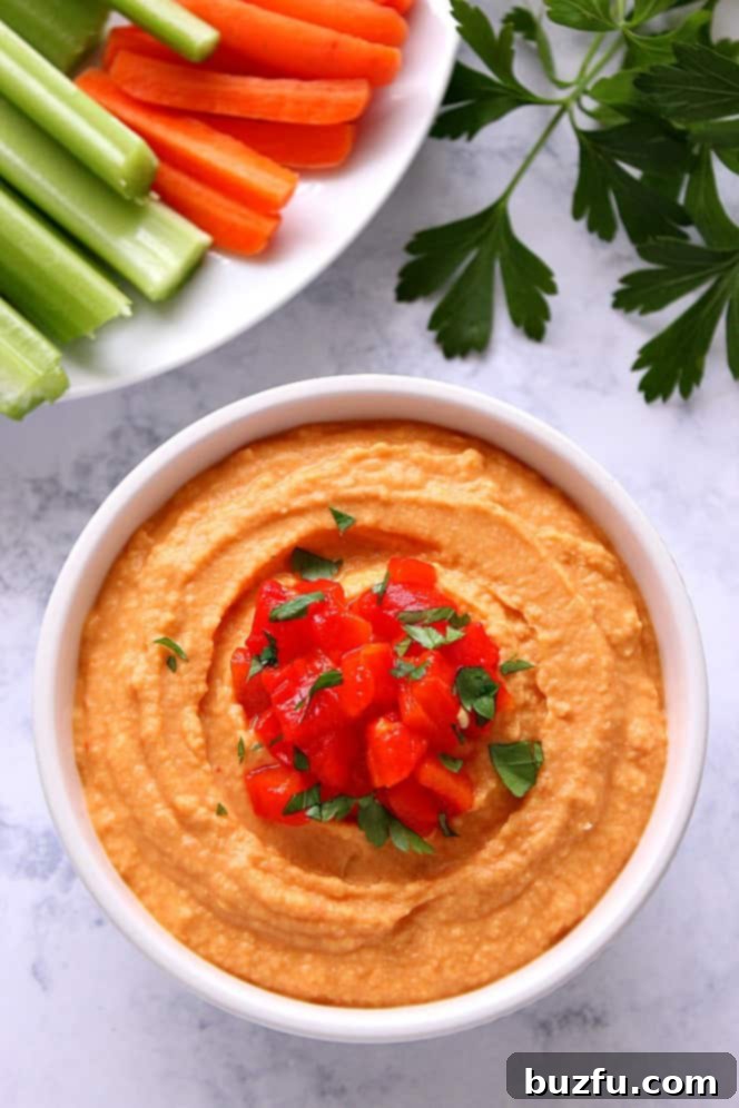 A beautifully presented bowl of Roasted Red Pepper Hummus on a marble surface, surrounded by crisp veggie sticks and fresh parsley, ready for a healthy snack.