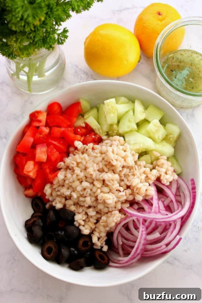 A large mixing bowl filled with the base ingredients for Greek salad: cooked barley, chopped cucumbers, thinly sliced red onion, ripe tomatoes, and briny olives.