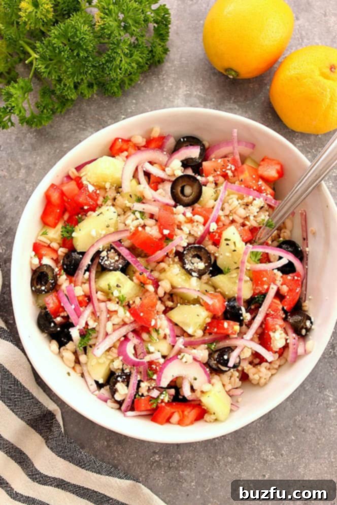 Close-up of a vibrant Mediterranean salad in a glass bowl, showcasing fresh ingredients like barley, tomatoes, cucumbers, red onion, and olives, coated in a light dressing.