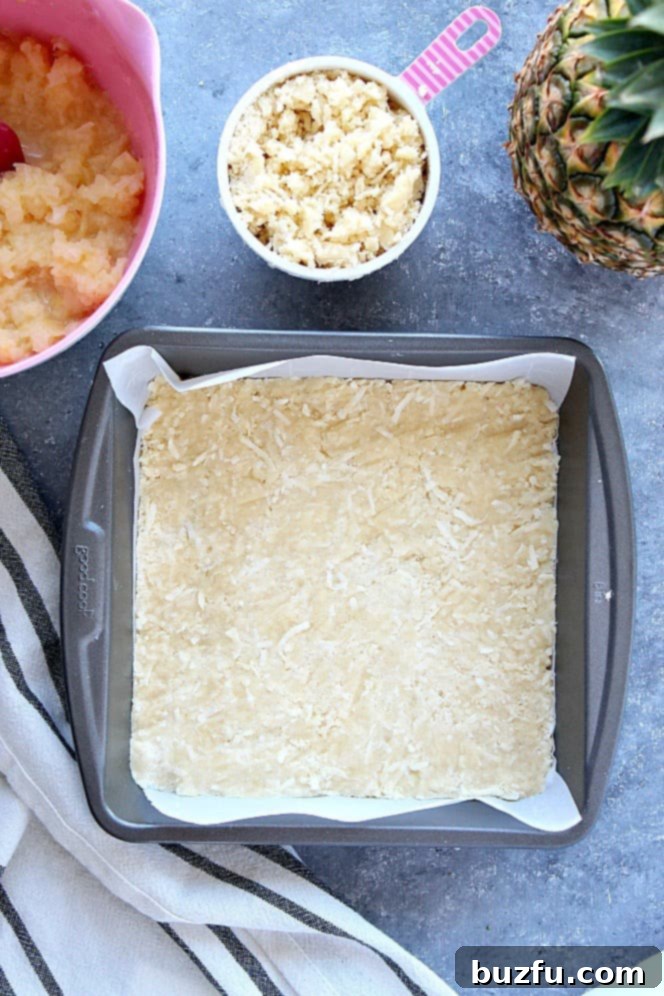The golden-brown crust of the tropical pineapple coconut crumb bars is pressed firmly into a square baking pan, showcasing its even texture, ready for the sweet pineapple filling.