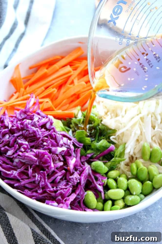 Close up on bowl with purple cabbage, carrots, green cabbage, edamame beans and dressing pouring from a glass cup. 