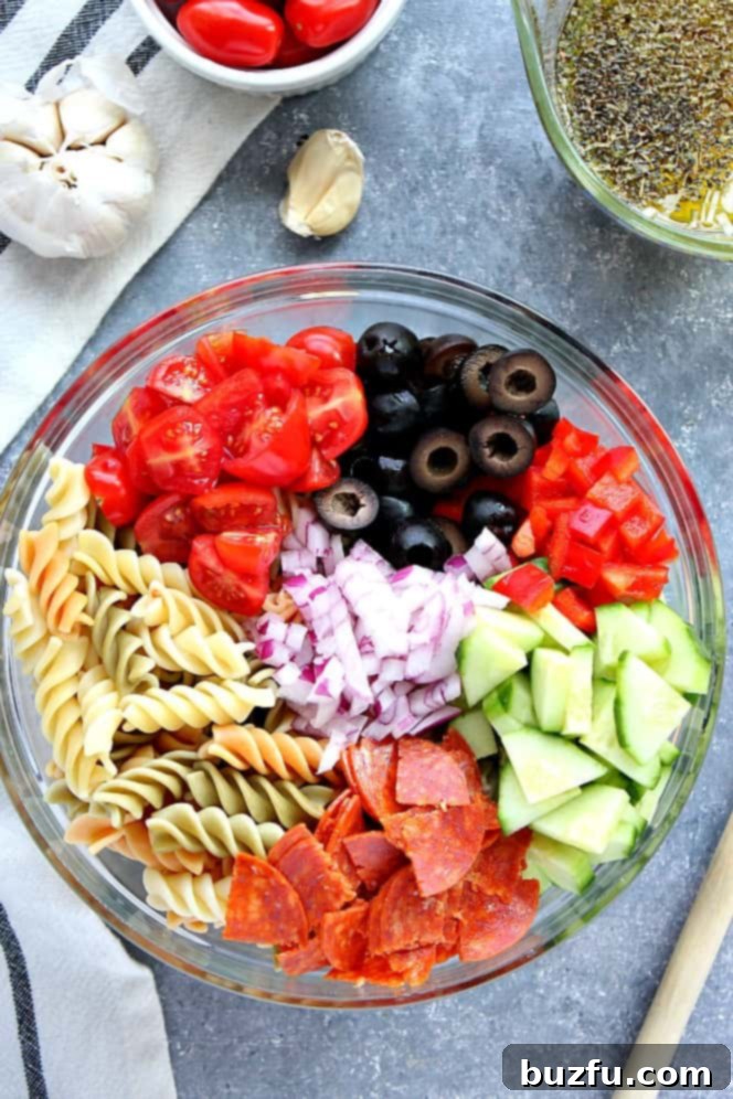 Overhead shot of pasta, tomatoes, olives, cucumber, pepperoni, red pepper and onion in a mixing bowl.