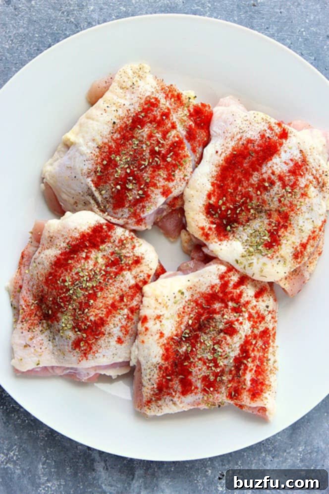 Overhead shot of raw bone-in skin-on chicken thighs, seasoned with salt, pepper, paprika and oregano, on white plate. 
