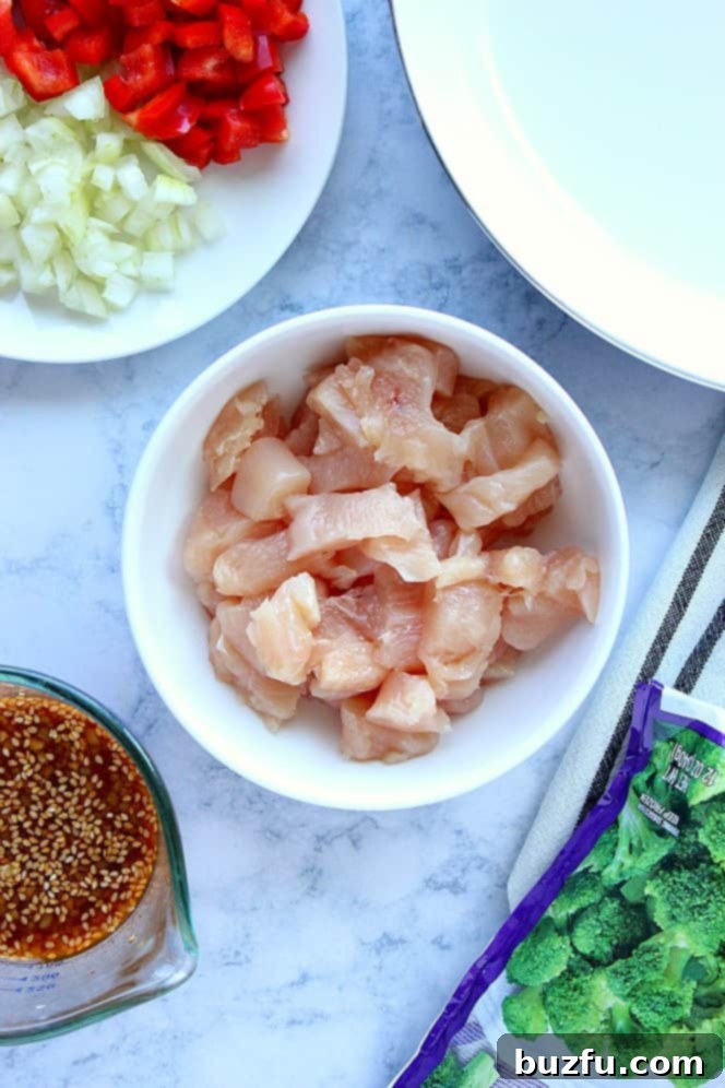 Overhead shot of all ingredients for Teriyaki Chicken Stir Fry: cubed chicken in a white bowl, sliced red bell pepper and onion on a plate, homemade teriyaki sauce in a cup, and frozen broccoli in a bag.