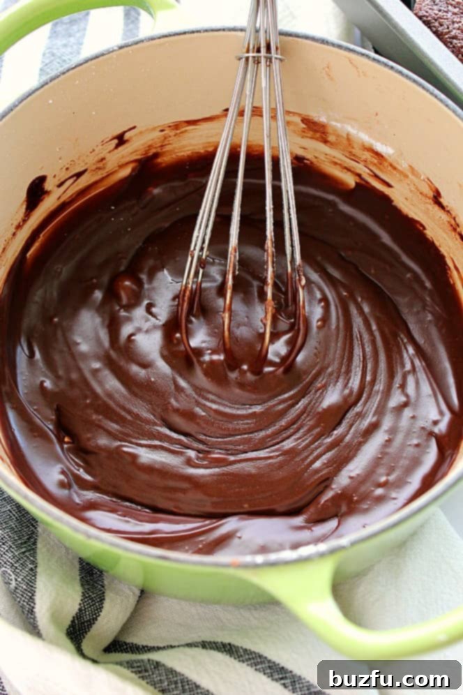 How to make fudgy frosting in a saucepan. Overhead shot of rich fudgy chocolate frosting in a saucepan, with a whisk resting on the side, demonstrating its smooth texture.