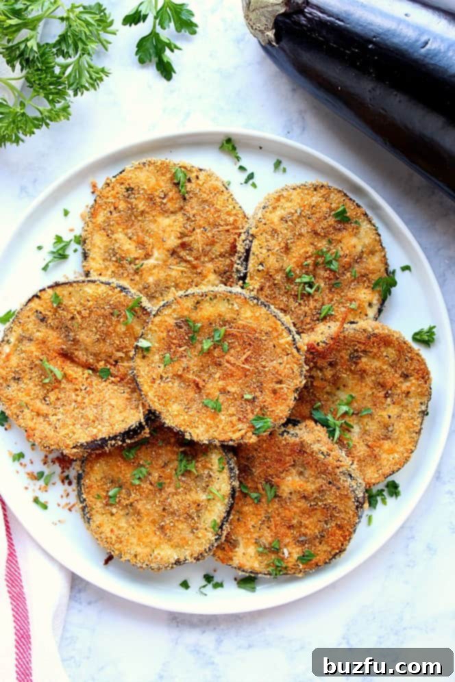 Garlic Parmesan Baked Eggplant Recipe - crispy baked, not fried, eggplant with garlic Parmesan breading. A delicious way to enjoy this fall vegetable! An inviting overhead shot of perfectly baked and crispy garlic parmesan eggplant, garnished with a sprinkle of fresh chopped parsley, presented on a white serving plate, ready to be savored.