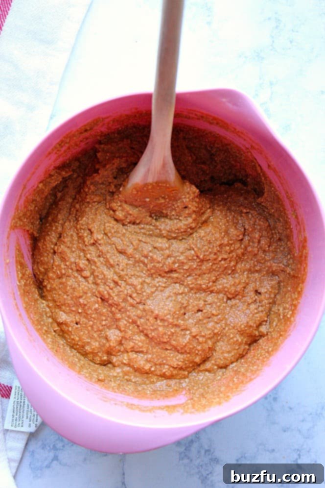 How to make gluten-free, vegan, healthy pumpkin muffins. Overhead shot of pumpkin muffin batter in a vibrant pink mixing bowl, with a wooden spoon resting inside. The batter is smooth and well-combined.