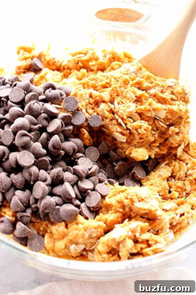 Close up shot of pumpkin oatmeal cookie dough in a glass bowl, with visible chocolate chips and a wooden spoon resting on the dough, showcasing its rich texture.