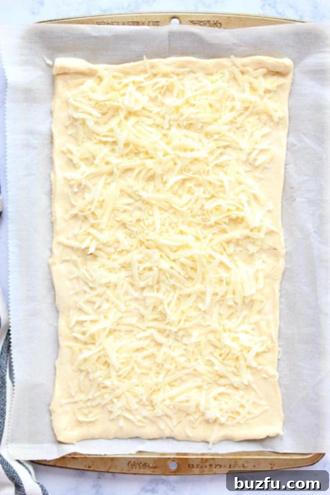 Overhead shot of crescent roll dough, neatly rolled out on parchment paper and generously topped with shredded cheese, ready for the next layer.
