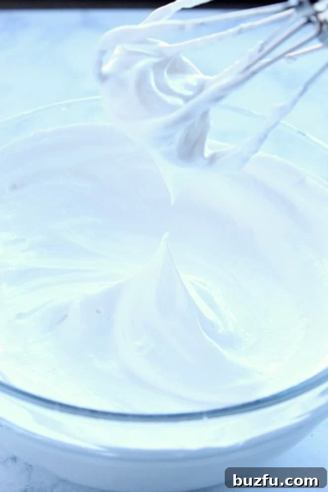 Close up shot of whipped egg whites with a stiff peak in the center and mixer beaters above bowl. 