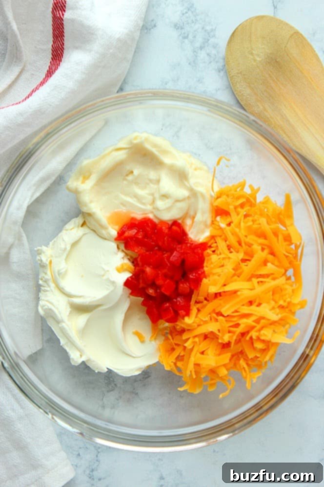 Overhead shot showcasing the primary ingredients for pimento cheese spread: vibrant shredded cheddar, creamy mayonnaise, soft cream cheese, and finely chopped pimento peppers, all in a glass mixing bowl.