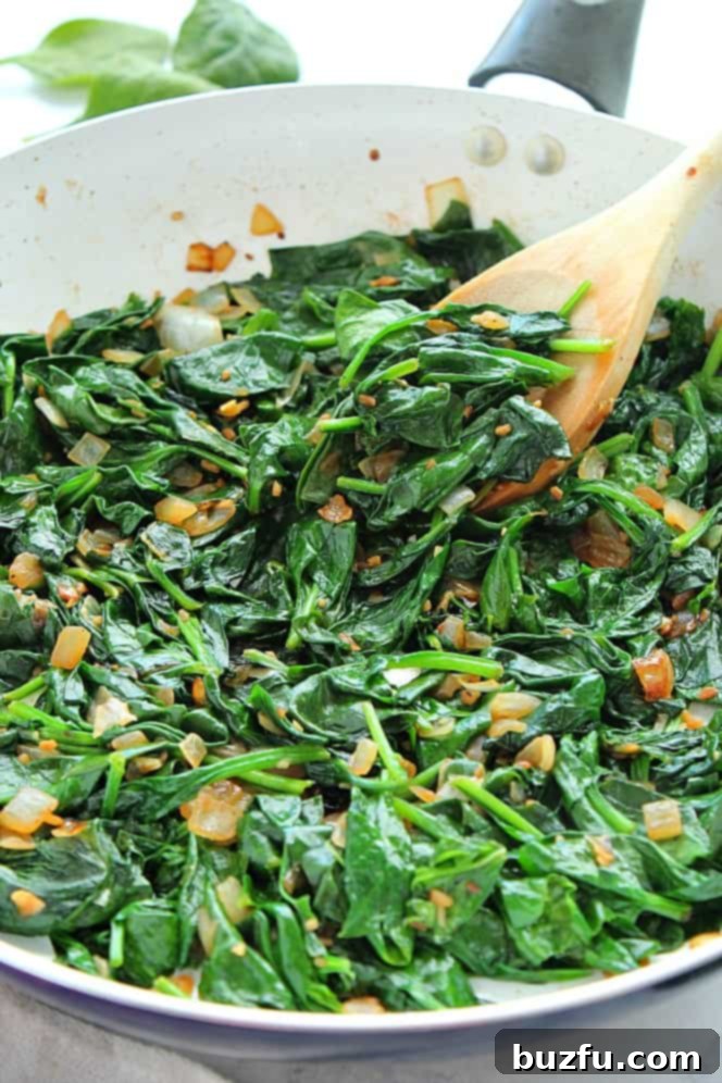 Side close-up shot of vibrant green sauteed spinach with caramelized onion in a large white pan, ready to serve.