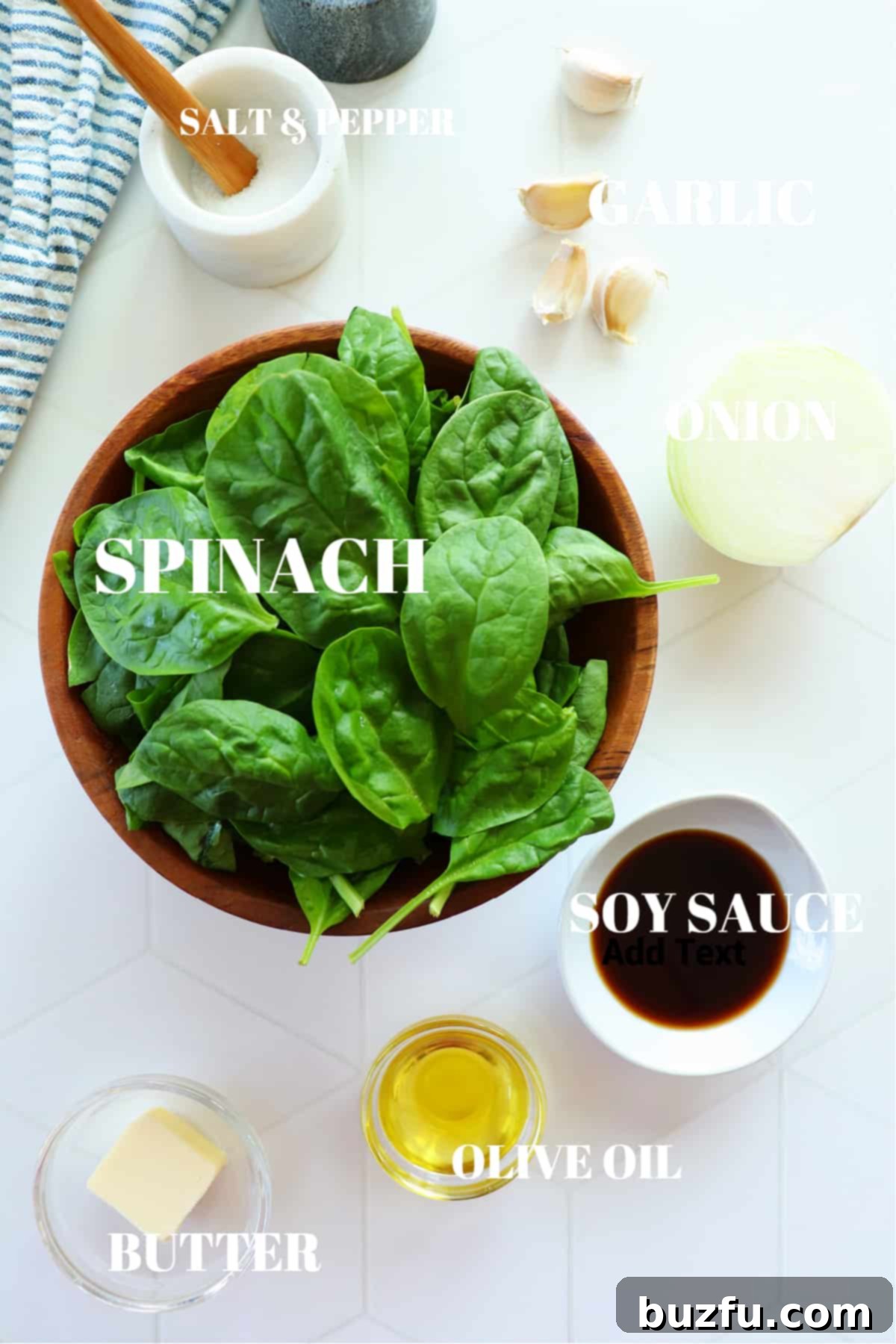 Bowls of fresh spinach, sliced onion, garlic cloves, a stick of butter, a bottle of olive oil, and a small dish of salt arranged on a white board.