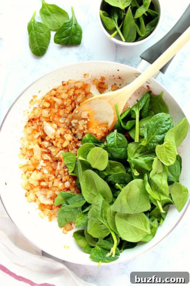 Overhead shot of diced onion and fresh green spinach leaves being tossed in a large skillet.