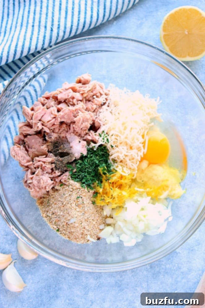 Overhead shot of tuna cakes ingredients: canned tuna, breadcrumbs, egg, cheese, lemon zest, onion and herbs in glass mixing bowl. All fresh and ready for preparation.