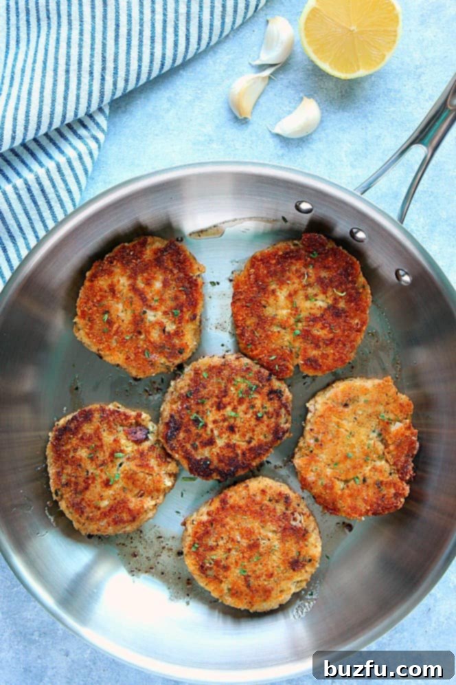 Overhead shot of 6 tuna cakes frying in a stainless steel pan, turning golden brown, with a striped kitchen towel in background.