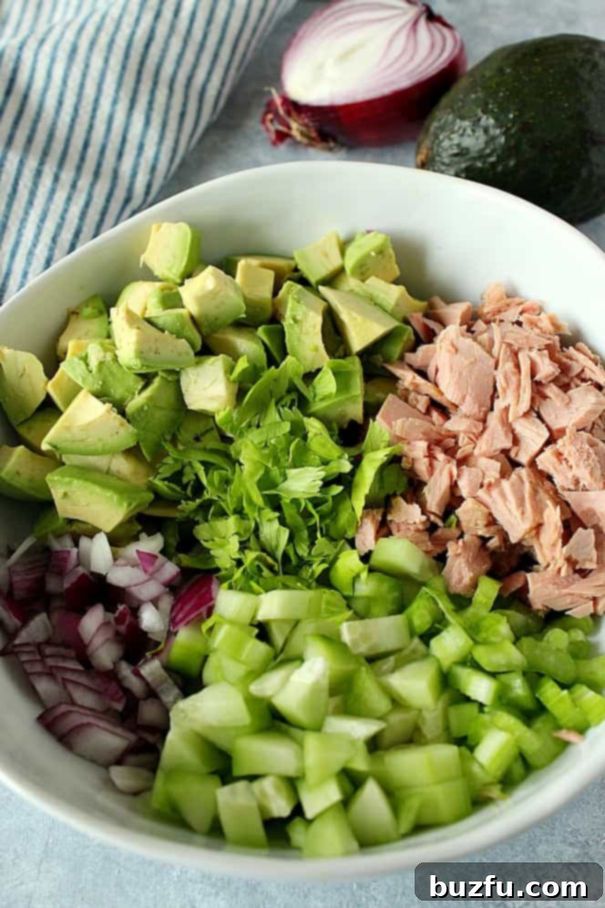 How to make avocado tuna salad step-by-step. Close-up side shot of fresh ingredients: tuna, avocado chunks, chopped celery, diced red onion, and cucumber slices, all arranged in a white salad bowl before mixing.