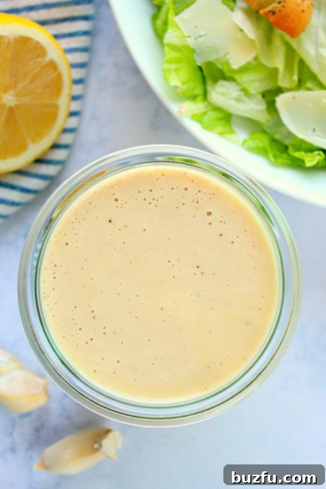 Overhead shot of creamy Caesar dressing in a glass jar, next to a bowl of fresh Caesar salad.