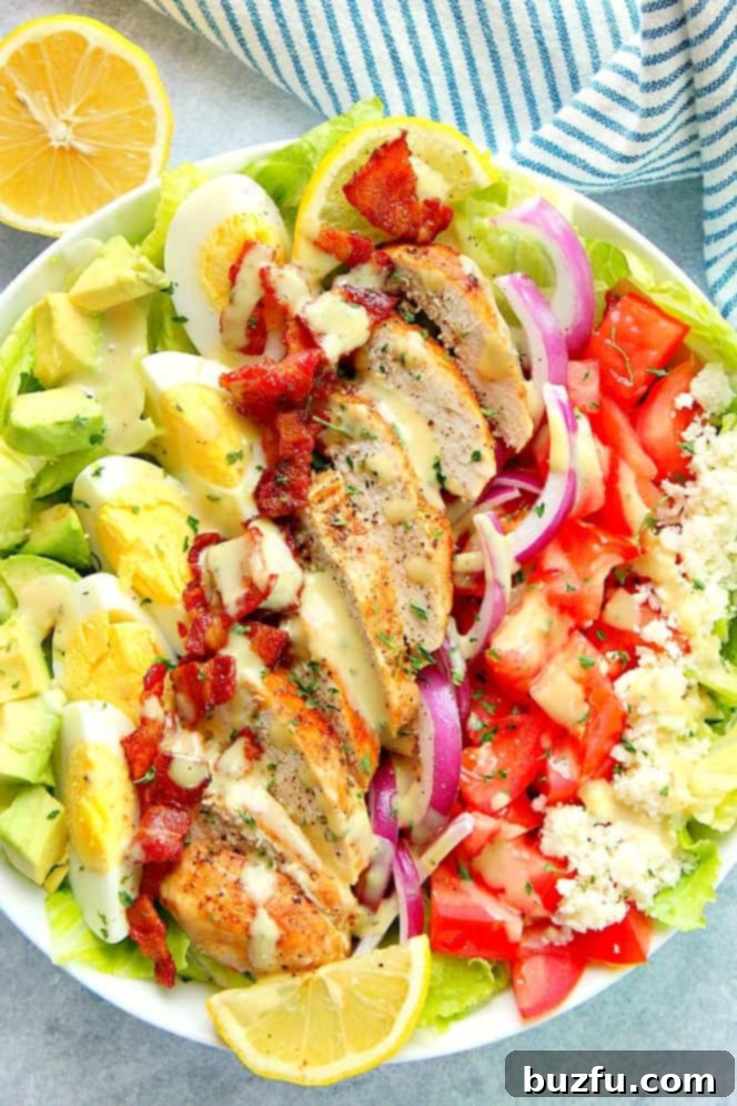 Chicken Cobb Salad with juicy lemon garlic chicken, crispy bacon, eggs, tomatoes, avocado, onion, lettuce and simple Dijon dressing. This whole-meal salad is perfect for lunch or dinner! Overhead shot of chicken Cobb salad in white salad bowl, showcasing its colorful ingredients like lemon garlic chicken, crispy bacon, eggs, tomatoes, avocado, and lettuce.