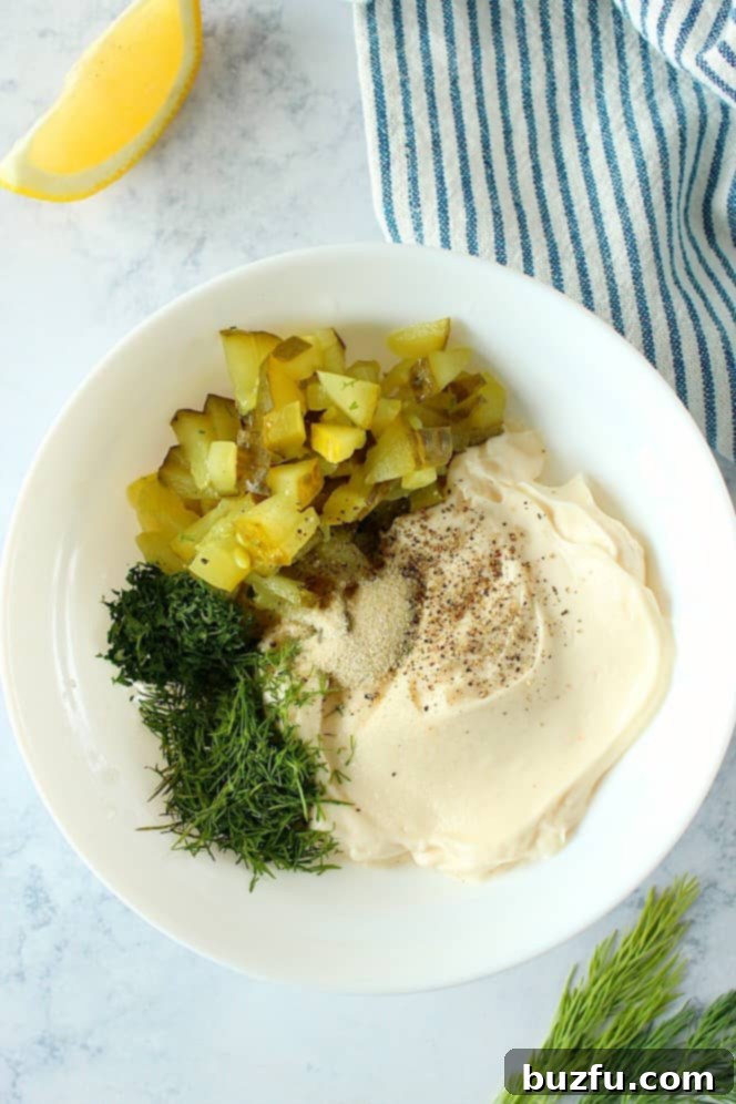 Overhead view of mayonnaise, chopped pickles, fresh dill, salt, and pepper in a white mixing bowl before combining.