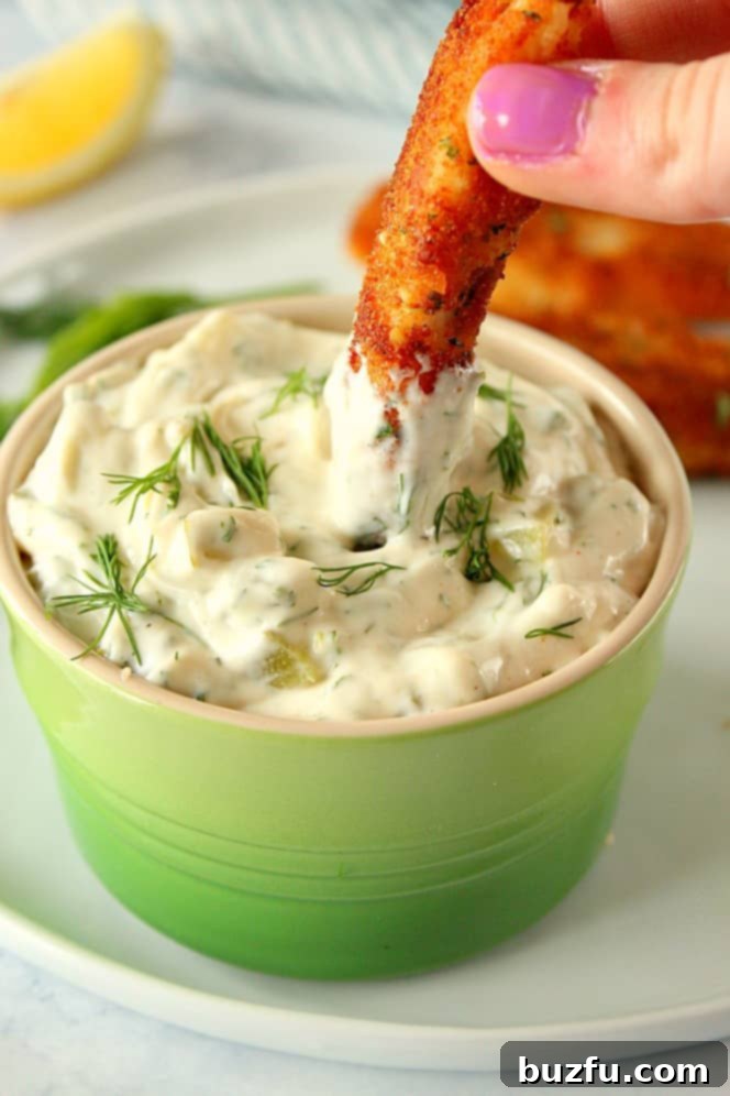 A golden, crispy fish stick being dipped into a bowl of homemade tartar sauce, highlighting its perfect texture.