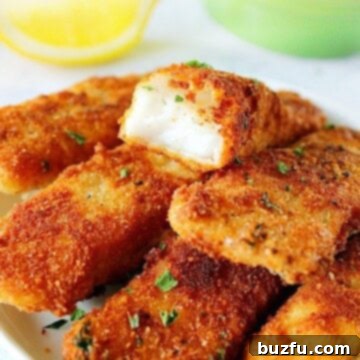 Golden Crispy Homemade Fish Sticks 2 Fried fish sticks stacked on a white plate with lemon wedges.