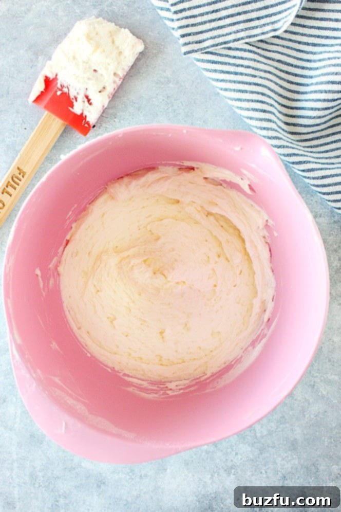 Achieving the perfect fluffy vanilla buttercream for your desserts. Overhead shot of creamy vanilla frosting in a pink mixing bowl, with more frosting on a spatula next to the bowl.