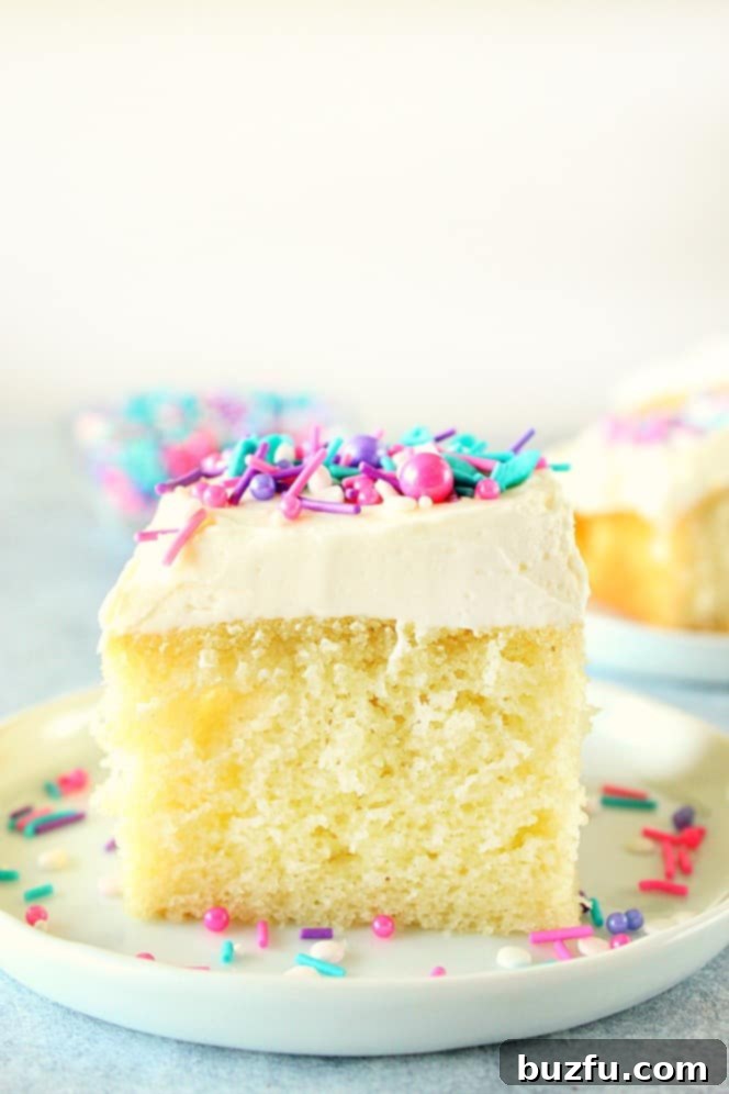 One Bowl Wonder Vanilla Cake 2 A perfectly square slice of vanilla cake, generously frosted with white vanilla buttercream and adorned with colorful sprinkles, rests on a pristine white plate. The cake shows its fluffy and moist interior texture, promising a delightful bite.