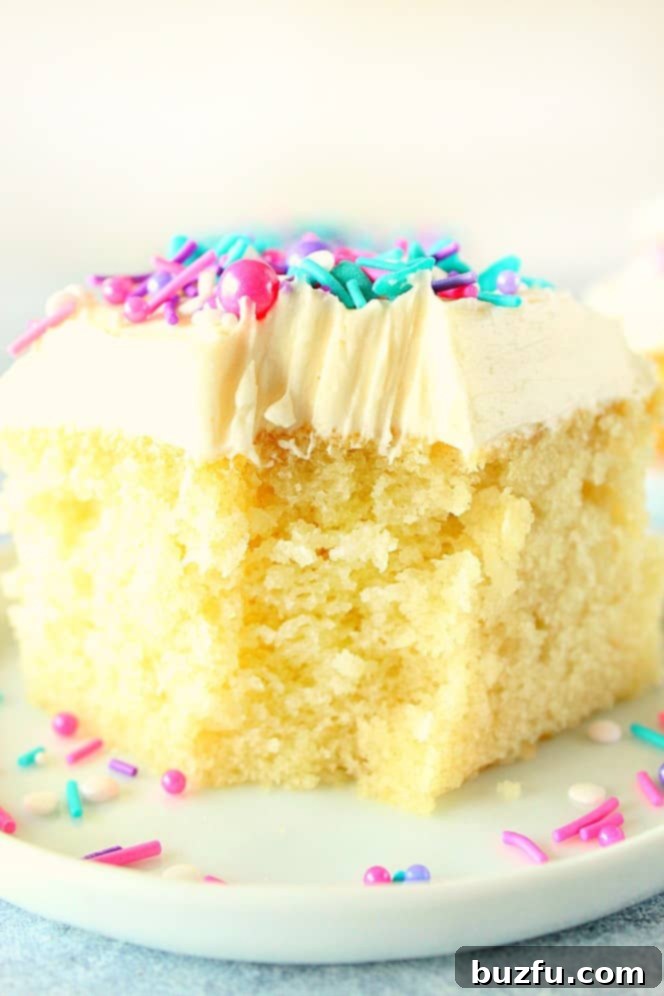 One Bowl Wonder Vanilla Cake 4 A close-up view of a slice of one-bowl vanilla cake, rich with fluffy vanilla frosting, sitting on a white plate. A bite has been taken from the front, showcasing its delicate, moist interior.