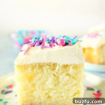 One Bowl Wonder Vanilla Cake 6 A perfectly square slice of vanilla cake, generously frosted with white vanilla buttercream and adorned with colorful sprinkles, rests on a pristine white plate. The cake shows its fluffy and moist interior texture, promising a delightful bite.