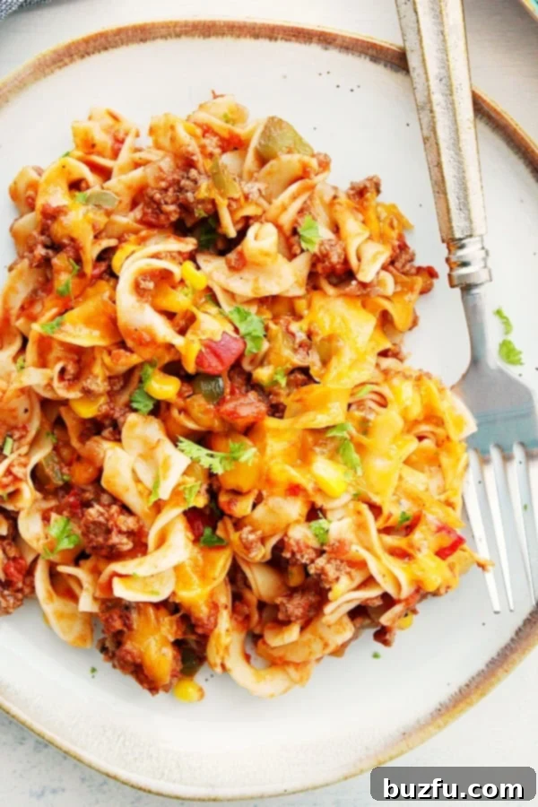 How to make the best ground beef casserole with egg noodles and tomato sauce. A serving of Cheesy Beef Noodle Casserole on a white plate, with a fork ready to dig in.