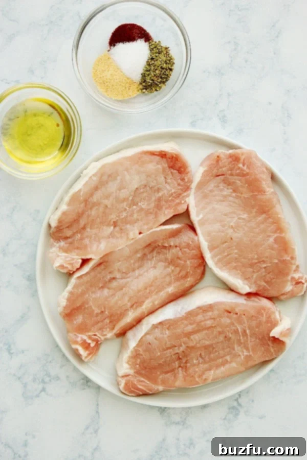 Herb-Crusted Baked Pork Chops 3 A collection of fresh ingredients for baked pork chops laid out on a marble cutting board, including pork chops, olive oil, and various spices.