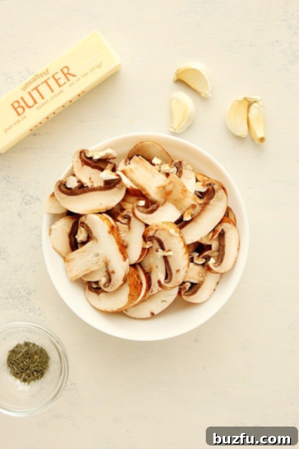 Fresh ingredients for sautéed mushrooms laid out on a white cutting board, including baby bella mushrooms, butter, garlic cloves, salt, pepper, and thyme.