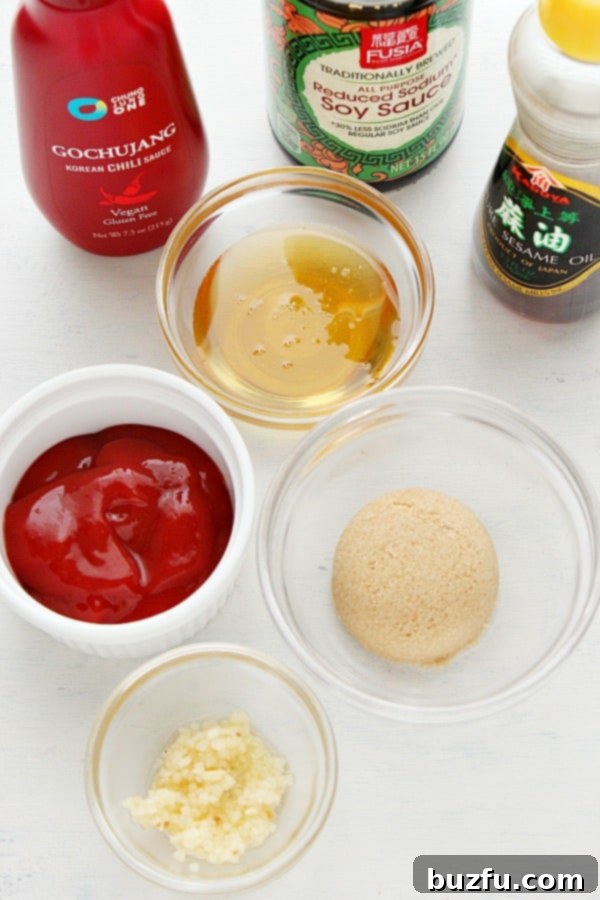 A collection of ingredients for Korean Gochujang sauce, including Gochujang paste, soy sauce, sesame oil, honey, brown sugar, ketchup, and garlic cloves, laid out on a wooden board.